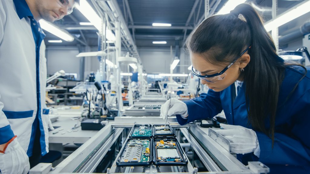  Two people in a factory, a man and a woman, are working on an assembly line. The woman is wearing a blue lab coat and safety glasses, and she is carefully working on a circuit board. The man is standing next to her and is watching her work.