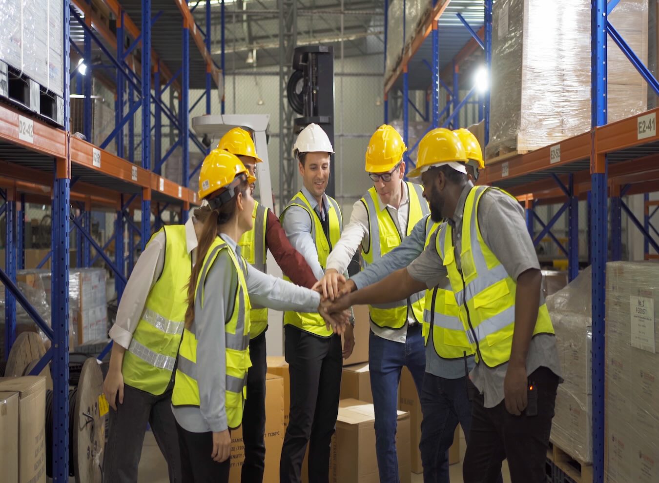 A group of warehouse workers wearing hard hats and vests shaking hands in a team setting, representing the collaboration and coordination needed to manage a large workforce in an industrial environment.