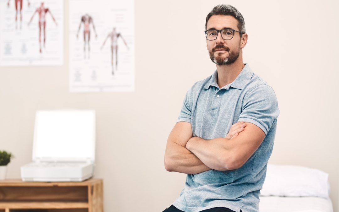 A man in casual dress sits in a doctor’s office with a neutral expression, waiting for an appointment, representing the importance of comprehensive employee benefits such as healthcare.